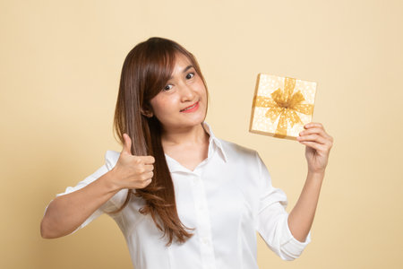 Young Asian woman thumbs up with a gift box on beige backgroundの写真素材