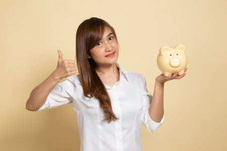Asian woman thumbs up with pig coin bank on beige backgroundの写真素材
