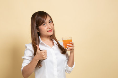 Young Asian woman thumbs up drink orange juice on beige backgroundの写真素材