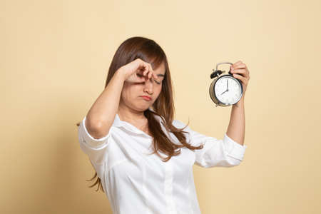 Sleepy young Asian woman with a clock in the morning on beige backgroundの写真素材