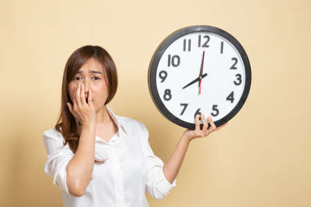 Sleepy young Asian woman with a clock in the morning on beige backgroundの写真素材