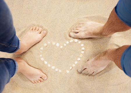 Female and male feet closeup of woman and man standing next to the shell heart at the sandy beachの写真素材