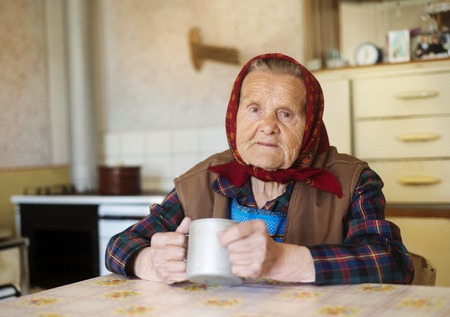Very old woman in head scarf is drinking tea in her old country style kitchenの写真素材