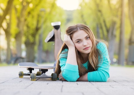 Portrait of beautiful teenage girl with skateboard lying on pavement in green alleyの写真素材
