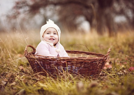 Happy little girl in warm clothes is sitting in basket in autumn natureの写真素材