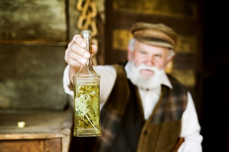 Portrait of old farmer with beard and hat holding a bottle of herbal spirit