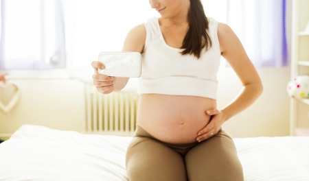 Beautiful pregnant woman taking selfie with mobile phone in her bedroom