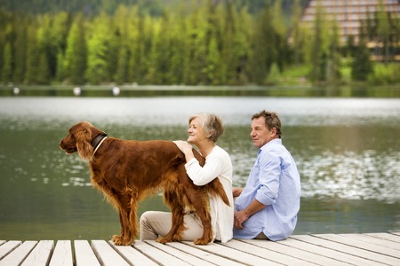 Senior couple with dog sitting on pier above the mountain lake with mountains in backgroundの写真素材