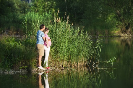Happy young pregnant couple hugging in nature, reflecting in the lakeの写真素材