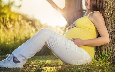 Outdoor portrait of beautiful pregnant woman sitting by the tree and holding her bellyの写真素材