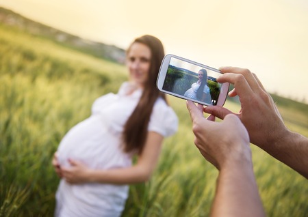 Man taking picture with smartphone of beautiful pregnant woman in the fieldの写真素材