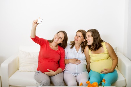 Three pregnant women sitting on sofa, smiling and taking selfie