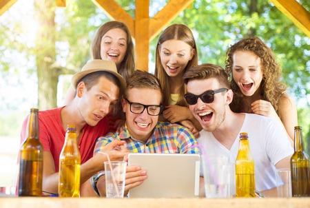 Group of happy friends drinking and having fun with tablet in pub garden