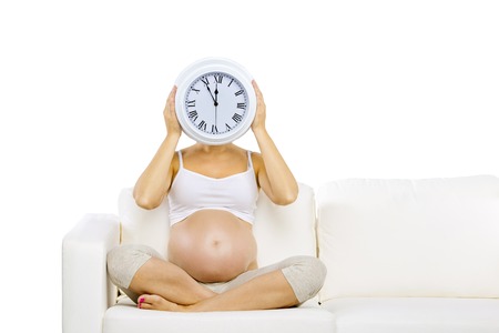 Studio portrait of pregnant woman holding big wall clock sitting on sofa isolated on white backgroundの写真素材