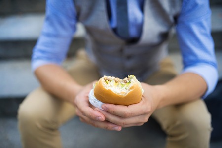 Deatil of modern hipster businessman having lunch break, sitting on stairs in town center and eating burgerの写真素材
