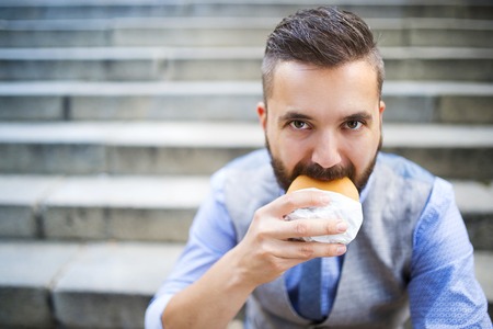 Modern hipster businessman having lunch break, sitting on stairs in town center and eating burgerの写真素材
