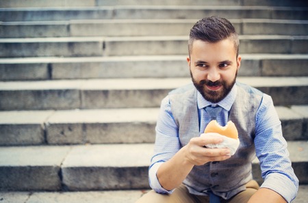 Modern hipster businessman having lunch break, sitting on stairs in town center and eating burgerの写真素材