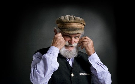 Portrait of old bearded man in traditional cap holding his moustache, posing in studio on black...