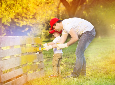 Cute little boy and his father in red caps painting wooden fence together on sunny day in natureの写真素材