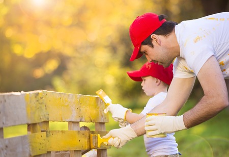 Cute little boy and his father in red caps painting wooden fence together on sunny day in natureの写真素材