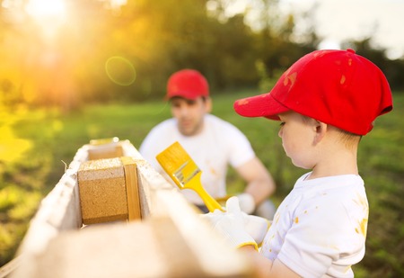 Cute little boy and his father in red caps painting wooden fence together on sunny day in natureの写真素材