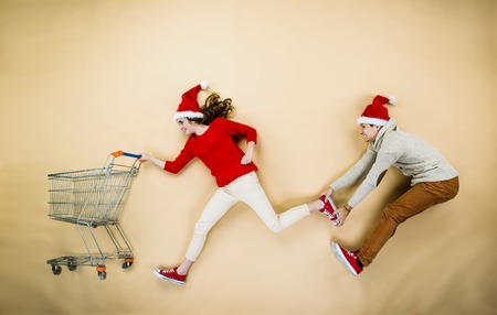Young couple in Christmas hats having fun running with shopping trolley against the beige backgroundの写真素材
