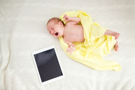 Crying newborn baby girl lying on bed next to digital tabletの写真素材