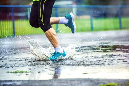 Young man running on asphalt sports field in rainy weather. Details of legs and sports shoes splashing in puddles.の写真素材