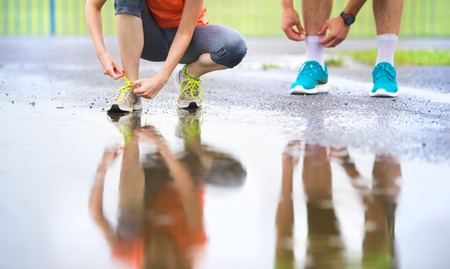 Young couple tying laces on asphalt sports field in rainy weather. Details of legs and sports shoes.