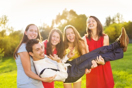 Funny portrait of bridesmaids holding groom on their hands in green sunny parkの写真素材