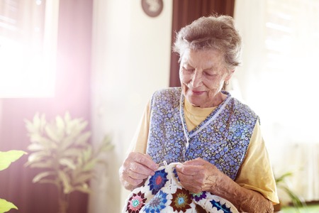 Old woman is knitting a blanket inside in her living roomの写真素材