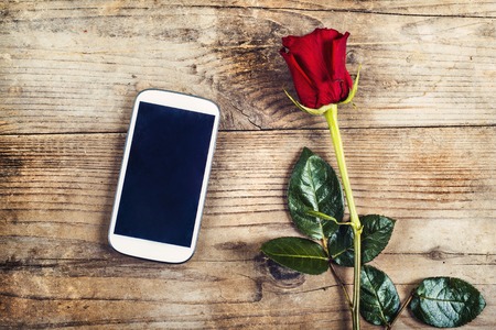 Valentine\'s day composition of yet not written love message in a phone and a red rose. Studio shot on a wooden floor backround.の写真素材