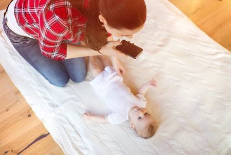 Young mother taking a picture of her cute little baby girl lying on a blanket in a living room.