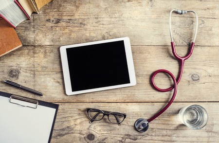 Workplace of a doctor. Tablet, stethoscope, clip board, books, glasses and other things on wooden desk background.の写真素材