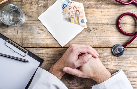 Hands of unrecognizable doctor accepting a bribe. Wooden desk background.の写真素材
