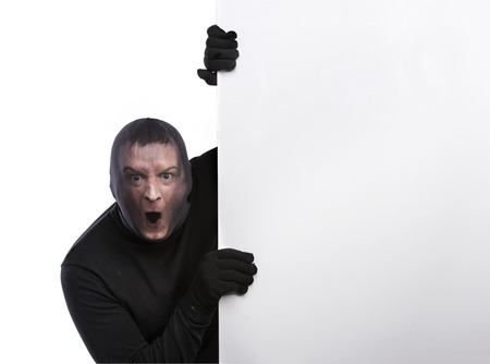 Thief with balaclava on his face, dressed in black, holding an empty banner. Studio shot on white...