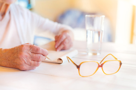 Hands of unrecognizable senior woman writing on a wooden white desk.の写真素材