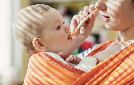 Young mother feeding her little daughter that she has in a baby carrier