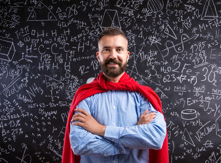 Hipster teacher in red cape standing against big blackboard with mathematical symbols and formulas. Studio shot on black background.の写真素材