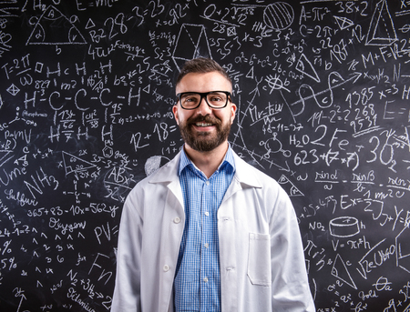 Close up of hipster teacher in white coat and eyeglasses against big blackboard with mathematical symbols and formulas. Studio shot on black background.の写真素材