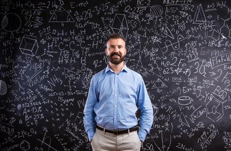 Hipster teacher standing against big blackboard with mathematical symbols and formulas. Studio shot on black background.の写真素材