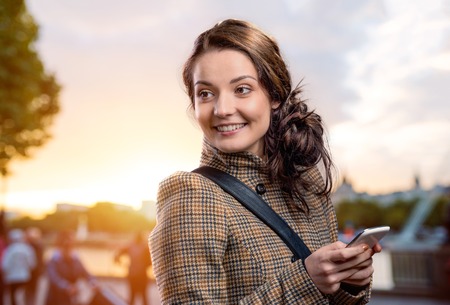 Young woman in checked brown winter coat with smart phone outside in sunny park