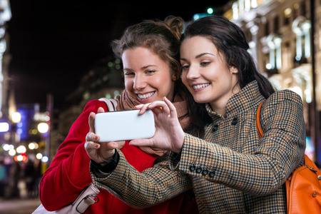 Two women in winter coats taking selfie with smart phone in the night city