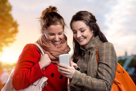 Two women in winter coats with smart phone outside in sunny park