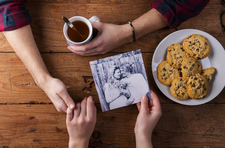 Unrecognizable man holding womans hand. Looking  at their black and white photo. Couple in love. Cookies and tea. Valentines day composition. Studio shot on brown wooden background.の写真素材