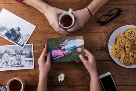 Unrecognizable mans and womans hand. Looking  at their black and white photos. Couple in love. Cookies and tea, daisy flower, smart phone. Valentines day composition. Studio shot on brown wooden background.の写真素材