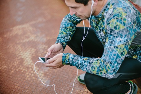 Runner in squat position holding smart phone and earphones, listening music, against old rusty metal bridgeの写真素材
