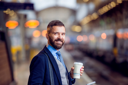 Hipster businessman holding a disposable coffee cup at the train station platformの写真素材