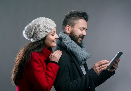 Woman in red winter coat and a hipster man with knitted scarf with tablet. Studio shot on gray background.の写真素材
