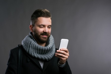 Young hipster man in black winter coat and knitted scarf holding a smartphone, texting. Studio shot...